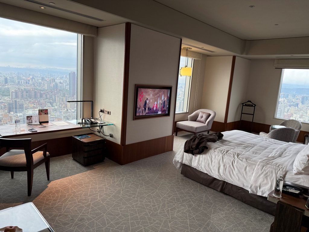 "Panoramic views from our room in Taipei, showcasing the city's skyline with the iconic Taipei 101 tower, a mix of modern and traditional buildings, and distant mountains under a cloudy sky."