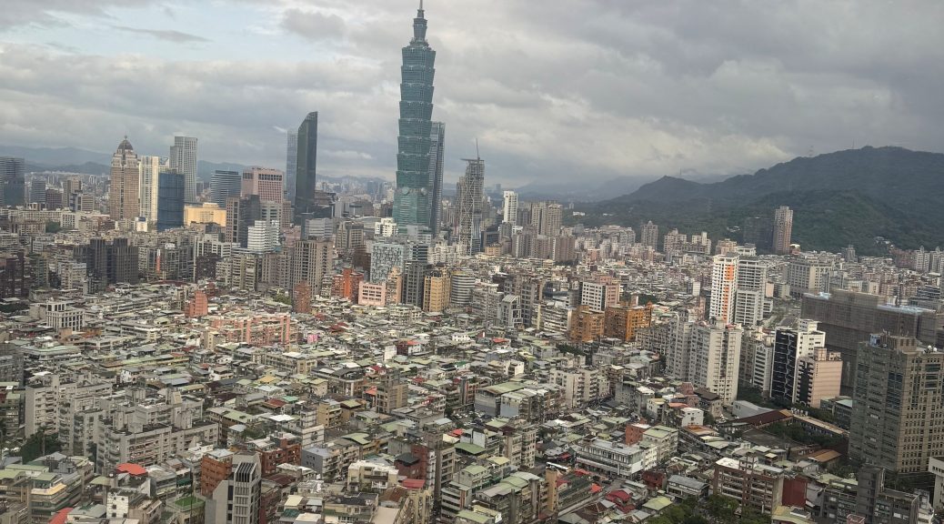 "Panoramic daytime view of Taipei city from the Shangri-La Far Eastern Hotel, featuring the iconic Taipei 101 skyscraper surrounded by a sprawling urban landscape and distant mountains under a cloudy sky."